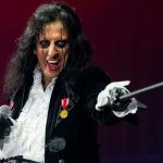 Alice Cooper performando no palco, usando roupas teatrais vitorianas, maquiagem marcante nos olhos, segurando um microfone e apontando uma bengala em direção ao público.