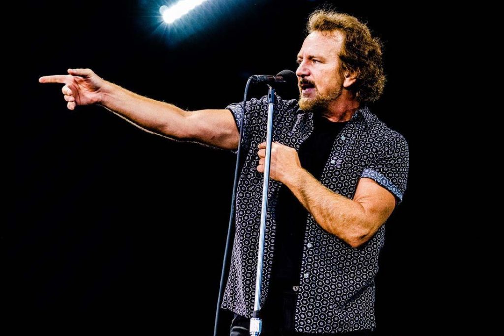 Eddie Vedder apontando para o público enquanto se apresenta ao vivo com microfone em mãos.