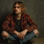 Pessoa jovem usando camisa xadrez e camiseta preta, em estilo grunge, sentada em frente a um muro pichado.