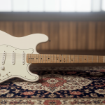 Guitarra Fender Stratocaster branca, com escudo branco e braço em maple, apoiada em um suporte sobre piso de madeira.