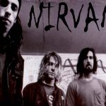 Foto em preto e branco dos integrantes do Nirvana — Kurt Cobain, Dave Grohl e Krist Novoselic — em frente a uma parede pichada, representando a estética sombria e autêntica do movimento grunge.
