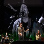 Integrantes da banda Pearl Jam se apresentando ao vivo com Eddie Vedder no centro, cantando com intensidade diante de uma grande projeção em preto e branco ao fundo.