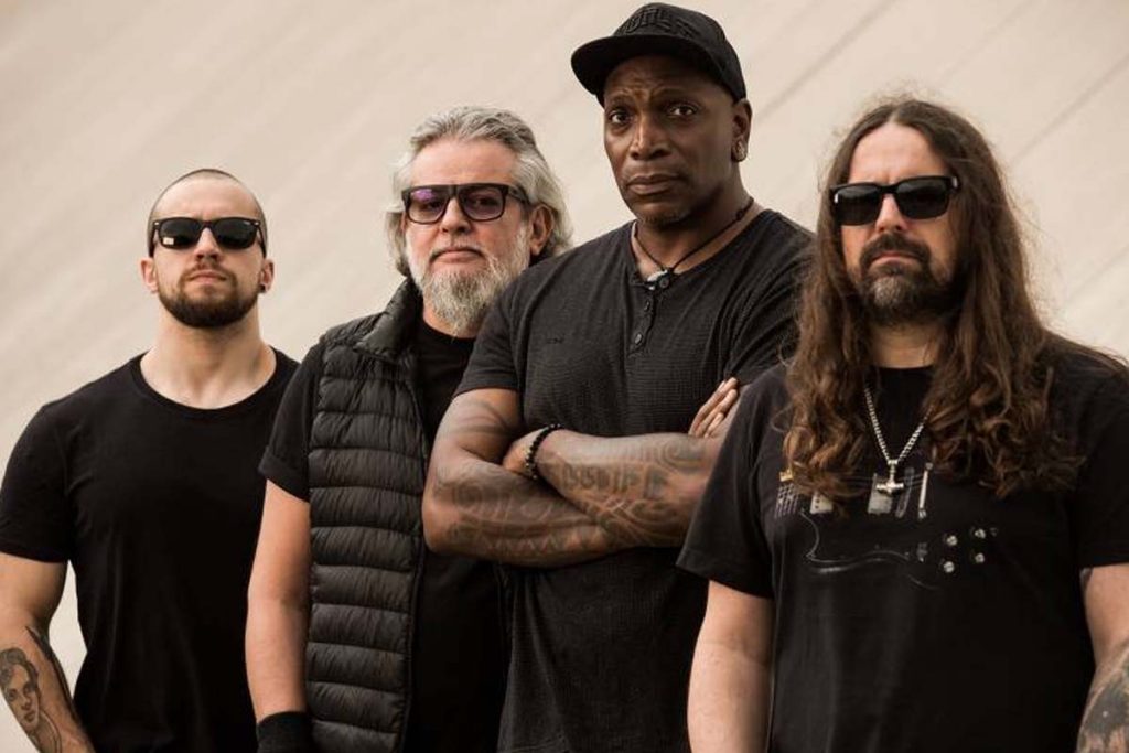 Quatro integrantes da banda Sepultura posam lado a lado, usando roupas escuras e óculos escuros, com expressão séria.